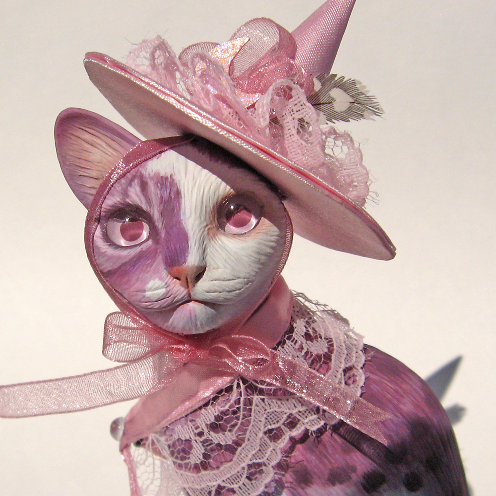 A handmade OOAK polymer clay sculpture of a pink witch kitty. She has pink eyes. Her precious face is half white. The pink hat she wears is made from satin fabric.