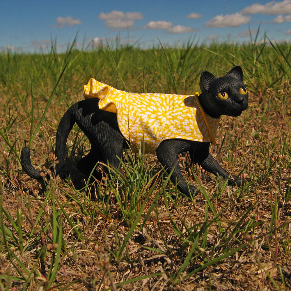 A handmade resin feline fashion doll wearing a stunning vintage yellow dress. She is prowling through green grass under a blue summer sky. Her bright yellow dress contrasts with her black fur in a striking way.